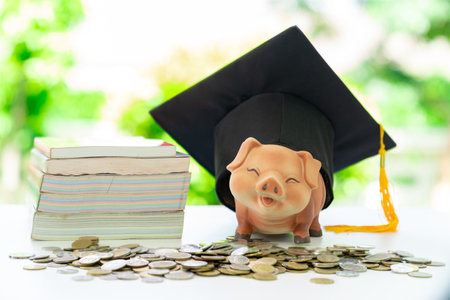 A graduation cap sits on top of a piggy bank, which is placed next to a stack of books and a pile of...