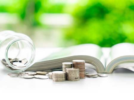 A jar of coins and an open book lie beside a pile of coins, representing the link between saving and learning.の写真素材
