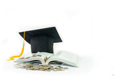 A graduation cap sits atop an open book, while a pile of coins lies in front it's symbolizing the cost and value of educationの写真素材