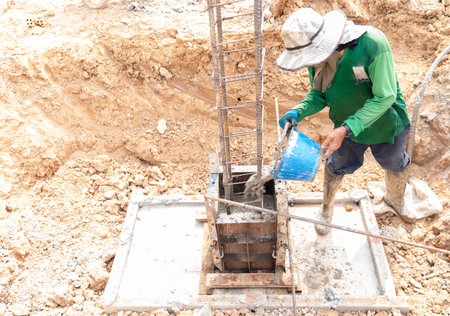 A construction worker is pouring concrete manually into a column foundation formwork using a blue...