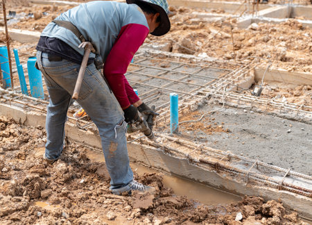 The worker wears gloves, jeans, and a wide-brimmed hat for sun protection. Visible rebar structure and blue PVC pipes are part of the foundation system.の写真素材