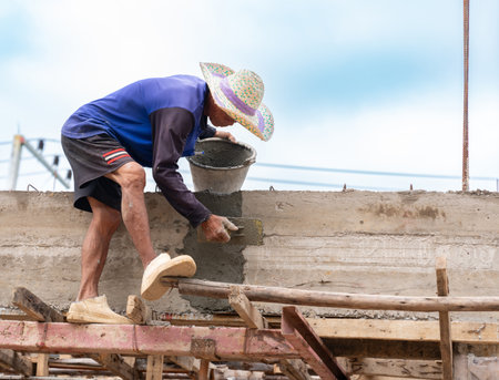 A construction worker, wearing a blue shirt, shorts, and a traditional straw hat, is applying cement or mortar to a concrete wall structure outdoors.の写真素材
