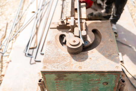 A worker wearing red gloves operates the rusty metal equipment to bend steel reinforcement bars, with other rebars and construction materials scattered nearby.の写真素材