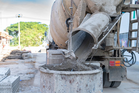 Wet concrete is poured from the rotating drum chute of a cement mixer truck into a large metal bucket or container on a sunny construction site.の写真素材