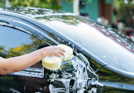 The man is focused on his task, demonstrating manual labor and self-care for his vehicle. In the background, there is a white house with dark windows, and a hose or water source is visible, confirming the setting as a residential or home garage/driveway area.の写真素材