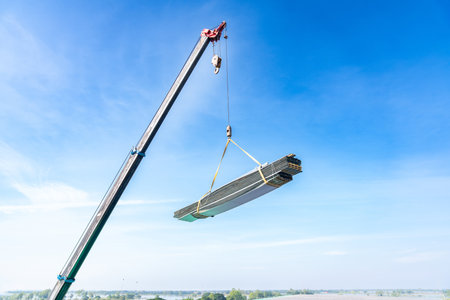 a mobile construction crane in operation, safely lifting a large, heavy bundle of metal profile sheets or roofing material high into the clear blue sky. The boom of the crane and the cables are prominently featured, demonstrating heavy equipment and modern logistics in construction.の写真素材