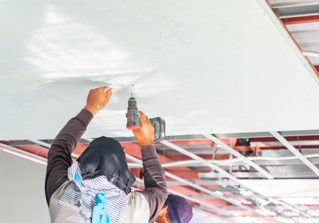 The worker is using a cordless electric drill to secure the panel, with his arms raised overhead. He is dressed in protective work attire, including a long-sleeve shirt and a covering over his head and neck for safety.の写真素材