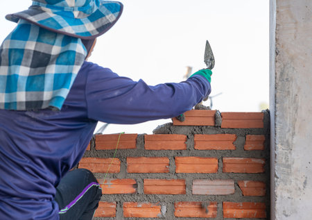 The worker's hands, wearing gloves, are applying fresh mortar with a trowel onto a course of red...