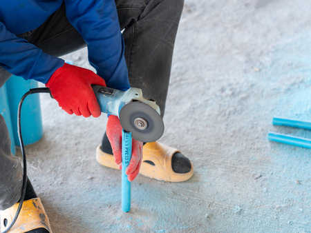 Close-up of Construction Worker Cutting Blue PVC Pipe with Electric Angle Grinder on Concrete Floorの写真素材