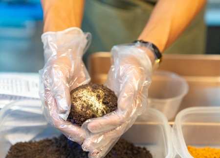 Close-up of a person wearing hygienic plastic gloves, manually kneading and coating a soft mochi ball with dark chocolate or cocoa cookie crumbles.の写真素材