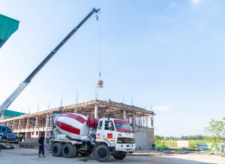 Construction workflow showing a cement mixer truck loading a crane bucket, which is then hoisted to workers who manually guide the wet concrete pour into building foundations at a rural site.の写真素材