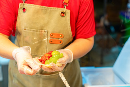 A close-up shot of a street food vendor's hands wearing hygienic gloves as they wrap fresh strawberries and green grapes into a soft mochi dough at a market.の写真素材