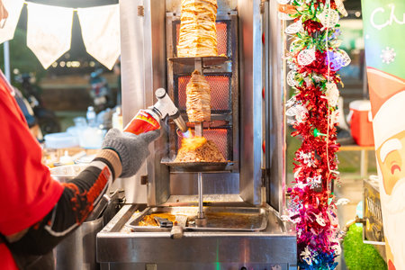 A street food vendor uses a handheld blowtorch to sear a vertical rotisserie of chicken kebab meat at a night market stall decorated with festive Christmas ornaments.の写真素材