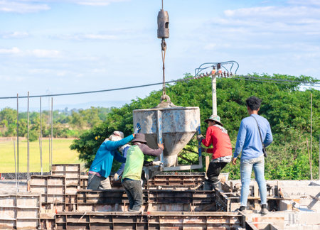 Construction workers manually guiding a large crane-suspended concrete bucket to pour wet cement into foundation formwork for a new building project in a rural landscape.の写真素材