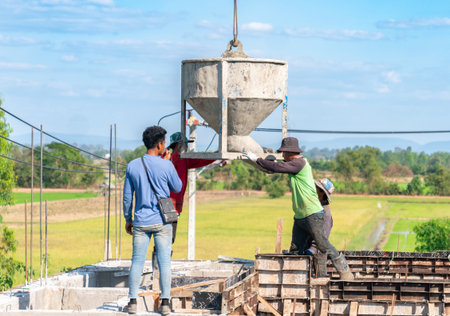 Construction workers manually guiding a large crane-lifted concrete bucket to pour wet cement into foundation formwork on a building site in a rural area.の写真素材