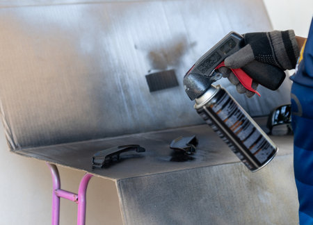 A person wearing protective gloves uses a black aerosol spray paint can with a trigger handle to recoat motorcycle parts on a cardboard-covered workspace.の写真素材