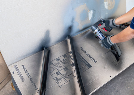 A person wearing protective gloves uses a black aerosol spray paint can with a trigger gun attachment to refinish and restore components on a cardboard-protected outdoor workspace.の写真素材
