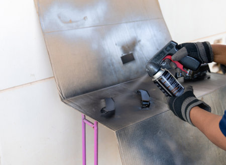 Close-up of a person wearing protective gloves using a black aerosol spray paint can with a trigger handle attachment to restore motorcycle parts on a cardboard-covered workspace.の写真素材