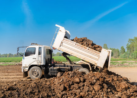 Heavy-duty tipper truck discharging a full load of raw earth and clay at a civil engineering site, preparing the foundation for new land development under a bright sky.の写真素材