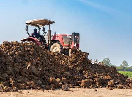 Red tractor with front blade leveling soil on a construction site or farm against a clear blue sky background.の写真素材