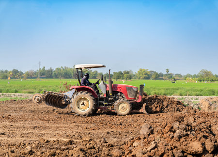 Red agricultural tractor with front dozer blade and disc harrow working on land preparation and soil leveling at a rural farm site under a clear blue sky.の写真素材