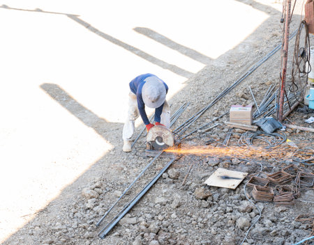 A high-angle view of a construction worker using a metal chop saw to cut steel rebar on a construction site with bright sparks flying across the ground.の写真素材