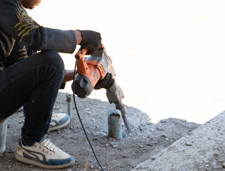 Professional construction worker using a heavy-duty electric demolition hammer to break concrete on a floor near a pipe at a building site.の写真素材
