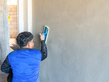 A professional construction worker in a blue uniform meticulously using a sponge float to create a smooth, even finish on a freshly plastered interior concrete wall.の写真素材