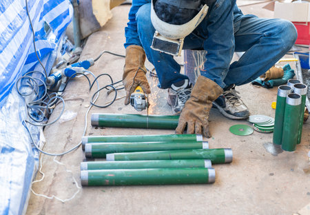 A metalworker wearing protective leather gloves and a welding mask uses a handheld power angle grinder to cut through a green steel pipe, creating bright orange sparks.の写真素材