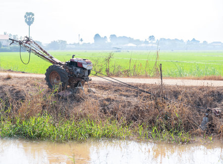 A two-wheel walking tractor, commonly used in Southeast Asian agriculture, sits parked on a dirt path next to an irrigation canal.の写真素材