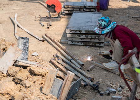 Industrial welder using an arc welding tool to join metal pieces on a dusty outdoor job site.の写真素材