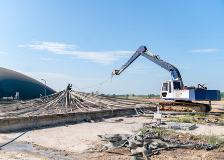 An excavator with a long-reach arm assisting a worker in securing a large industrial geomembrane or pond liner over a circular treatment tank.の写真素材