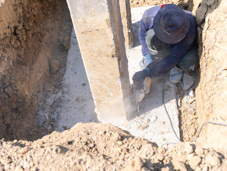 A laborer in a work hat uses an angle grinder to cut through a concrete foundation post in a deep trench at a construction development site.の写真素材