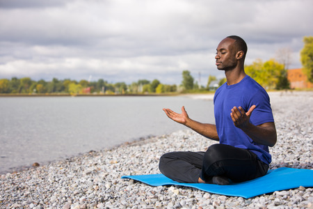 young black man wearing athletic wear sitting on the beach exercising yogaの写真素材