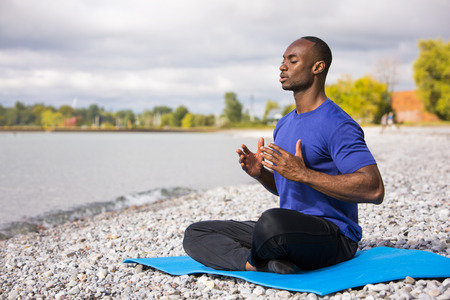 young black man wearing athletic wear sitting on the beach exercising yogaの写真素材