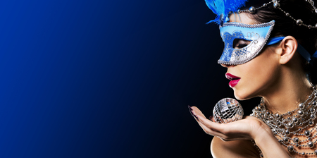 beautiful woman with dark makeup wearing blue and silver mask surrounded by party accessories on light black backgroundの写真素材