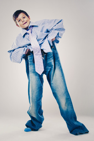 Caucasian boy wearing his Dad's shirt, jeans and tie on light background. He is wearing big adult size clothes which are too big for him.