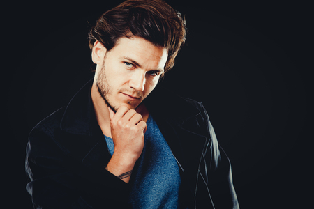 Close-up face of young bearded man on dark background modern and dramatic photo. Caucausian male model is wearing blue tshirt and black jacket.の写真素材