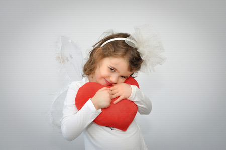 Cute young girl with a wings of fairy and alice band with a white bow holding a plush red heart on...