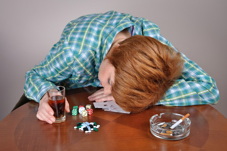 Exhausted woman with her head on the table sleeping next to cards, dice, pills and ashtray with cigarette holding a glass of alcoholの写真素材