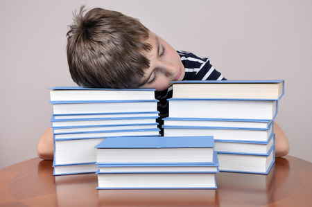 Tired young boy sitting and sleeping at the brown table with his head on the booksの写真素材
