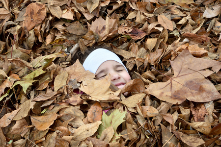 Smiling young girl with closed eyes covered with a heap of fallen autumn leavesの写真素材