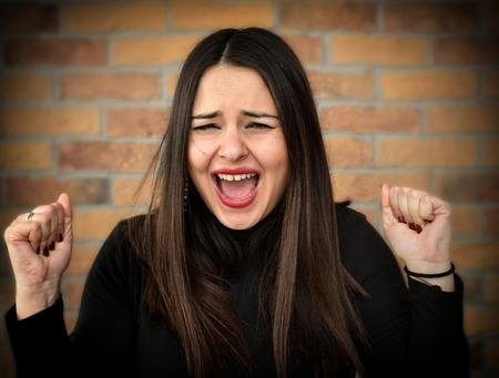 Beautiful young woman with clenched fist screaming in front of the brick wallの写真素材