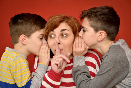 Sons whispering secret into ears of surprised mother with index finger on her mouthの写真素材