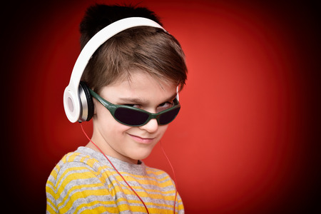 Young boy with headphones listening to music, enjoying and looking from under sunglasses. Isolated on the red background.の写真素材