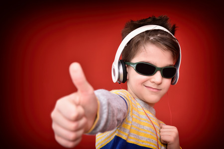 Young boy with headphones and sunglasses listening to music, enjoying and showing thumb up. Isolated on the red background. Selective focus.の写真素材