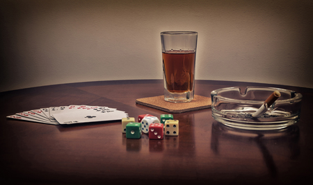 Glass of alcohol, cards, dice and ashtray with cigarette on the brown table. Filtered photo with toned and selective focus on the glass of alcohol drink.の写真素材