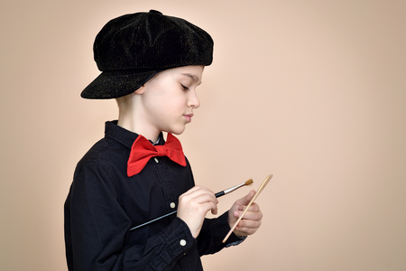 Cute young painter with red bow tie and black shirt and cap holding paint brush and art paletteの写真素材