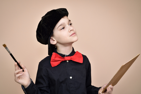 Thoughtful young painter with red bow tie and black shirt and cap holding paint brush and art paletteの写真素材