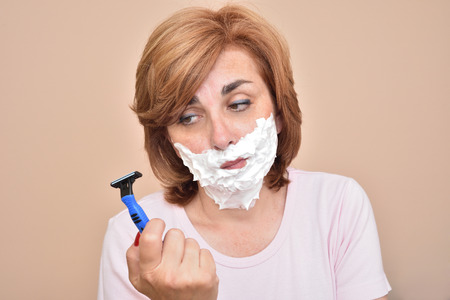Middle aged woman with shaving foam on her face holding and looking sadly at a razorの写真素材